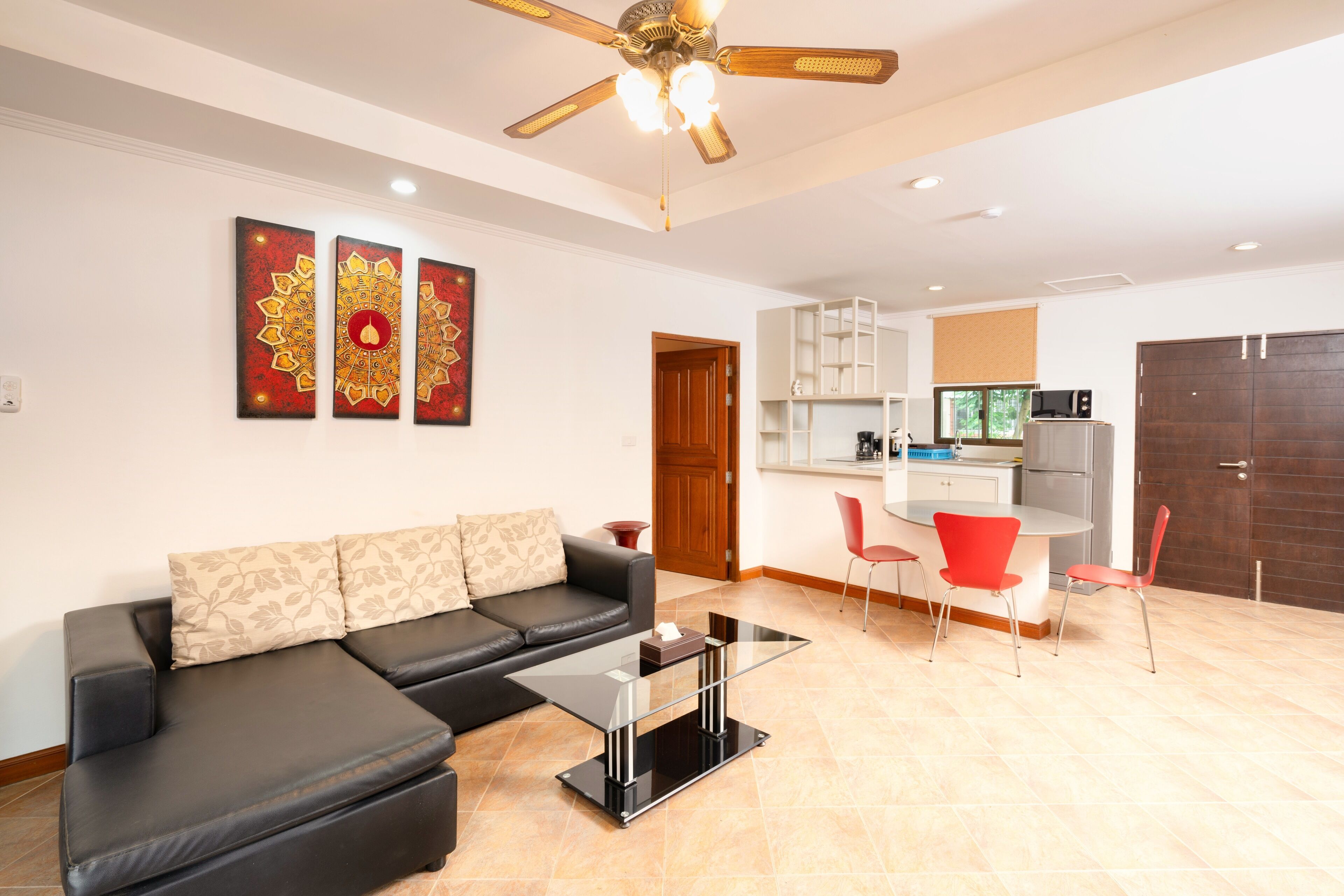 A modern living room with a black leather sofa, a glass coffee table, and a dining area with red chairs. There is a kitchen in the background with white cabinets and a microwave.