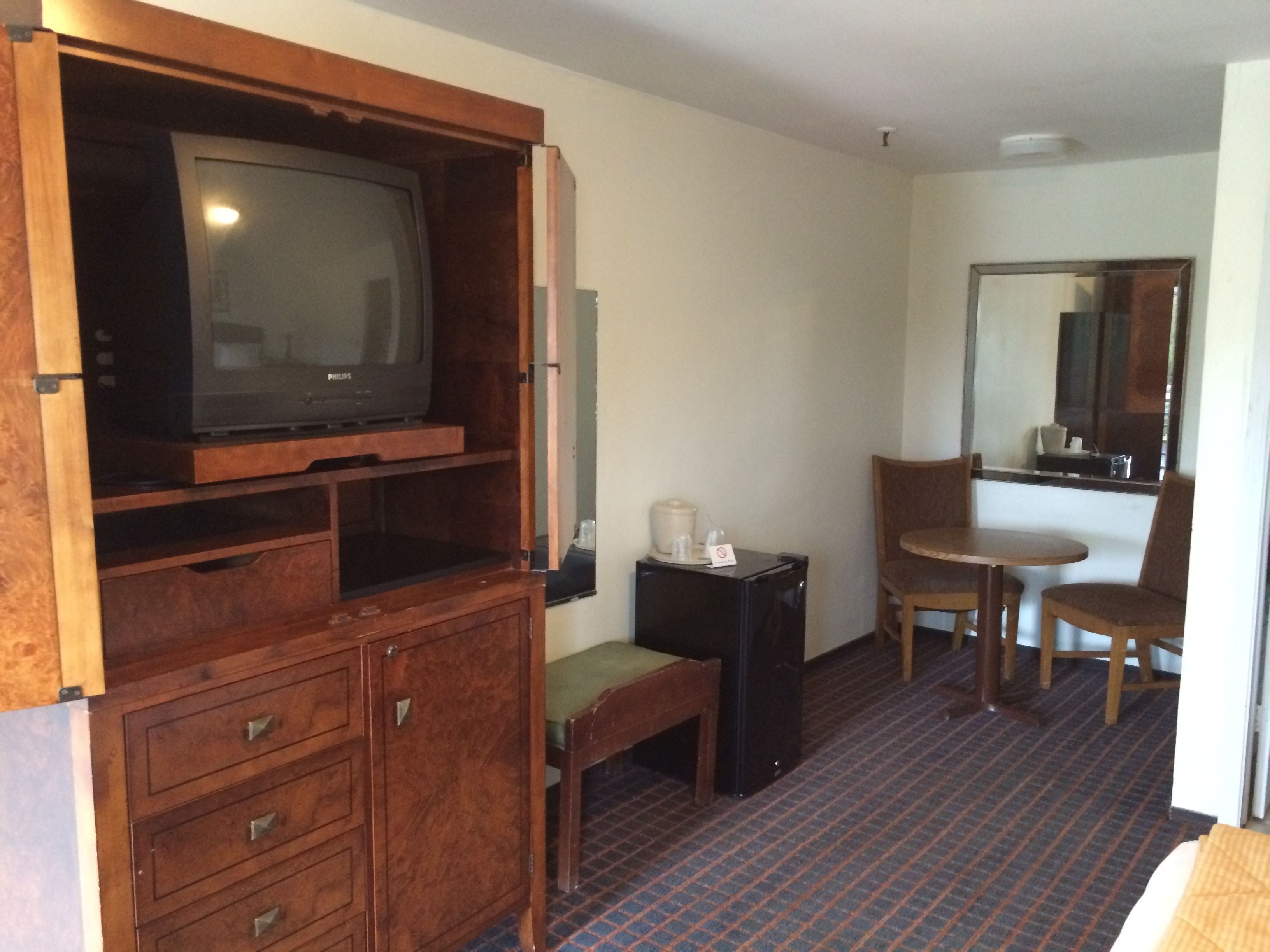 Une chambre d’hôtel comprenant un meuble de rangement en bois, une télévision, une petite table avec une chaise, un miroir et un lit.