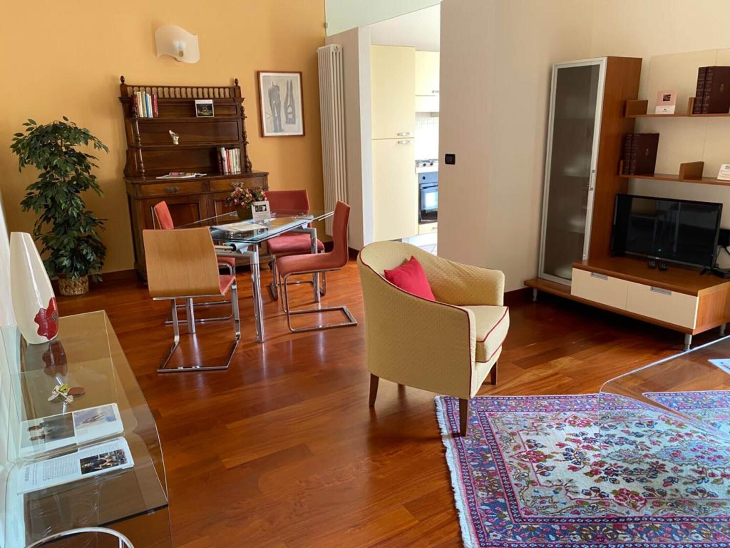 Una sala de estar con piso de madera, una mesa de centro de vidrio, un sofá color beige, un sillón rojo, una estantería y un televisor.