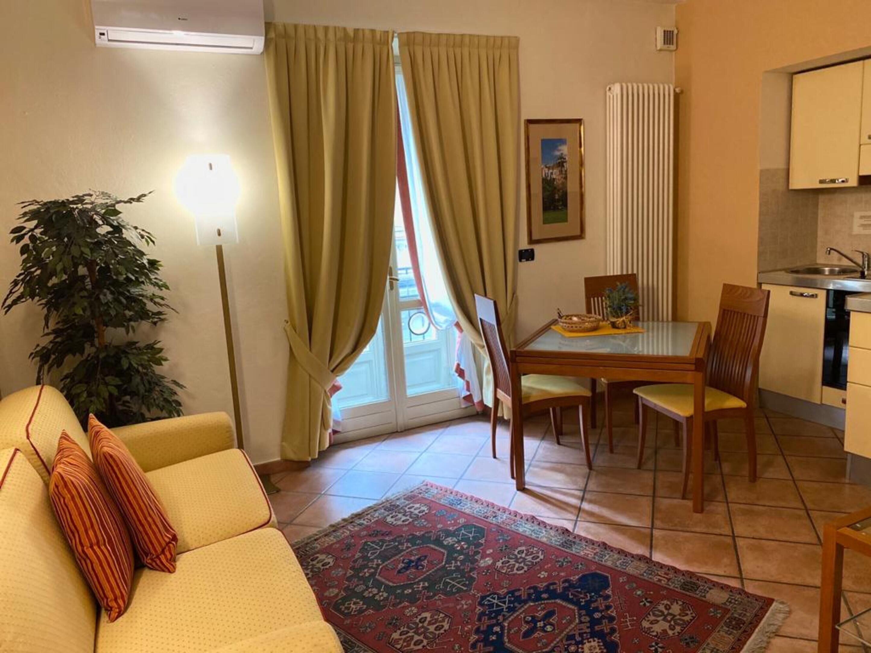 A living room with a sofa, a dining table with chairs, a potted plant, and a view of the outdoors through a window with curtains.