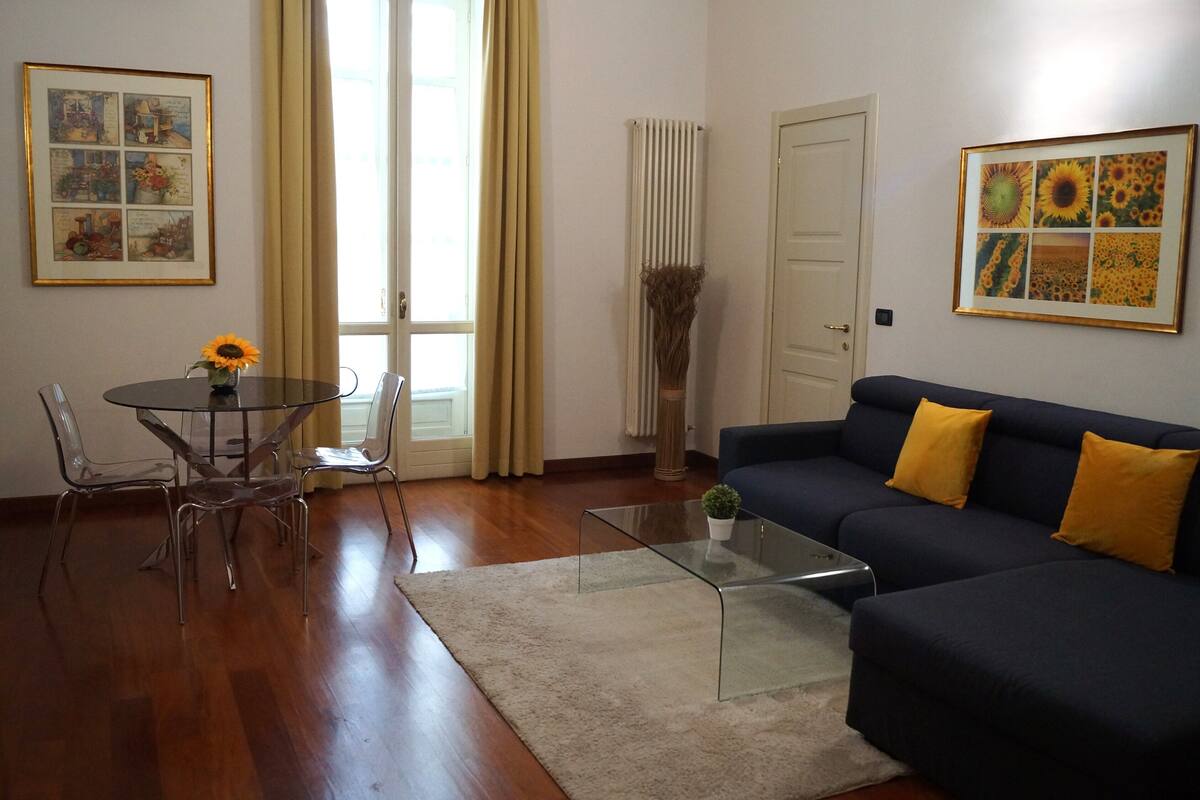A living room with a dark blue sofa, yellow cushions, a glass coffee table, and a dining area with a glass table and metal chairs.