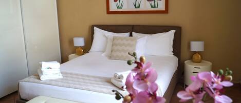 Quarto de hotel com uma cama grande, duas mesas de cabeceira com abajures, um banco no pé da cama e um quadro de flores na parede.