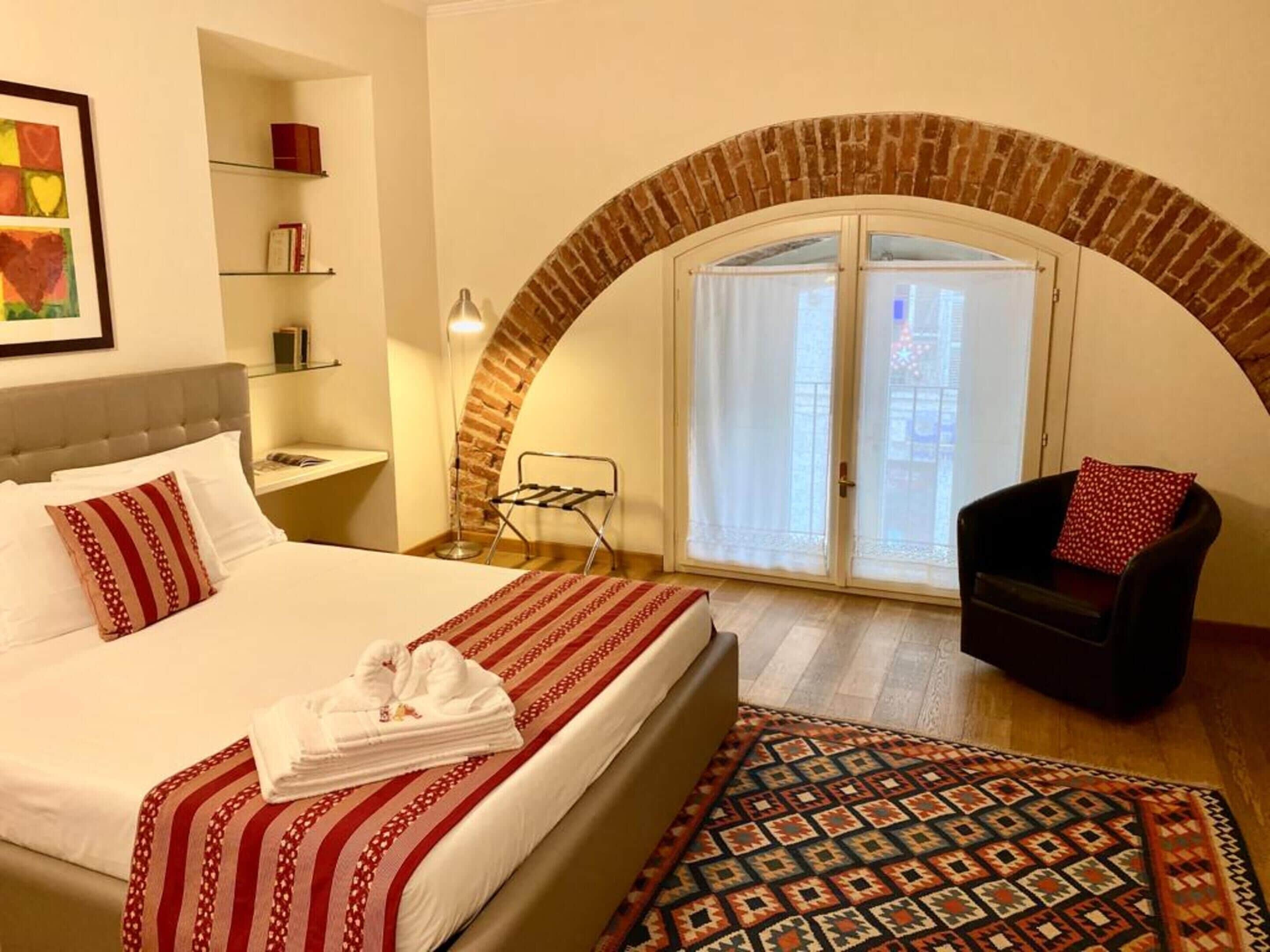Una habitación de hotel con una cama, una silla, una mesita, un estante con libros, un tendedero y una alfombra estampada.