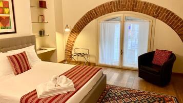 Una habitación de hotel con una cama, una silla, una mesita, un estante con libros, un tendedero y una alfombra estampada.