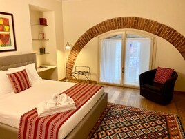 Quarto de hotel com cama, uma cadeira, uma mesinha, prateleira com livros, varal de roupa e um tapete estampado.
