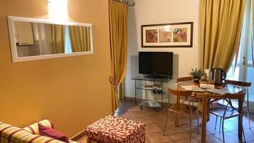 A living room with a sofa, ottoman, TV, glass table, chairs, curtains, and a framed picture on the wall.