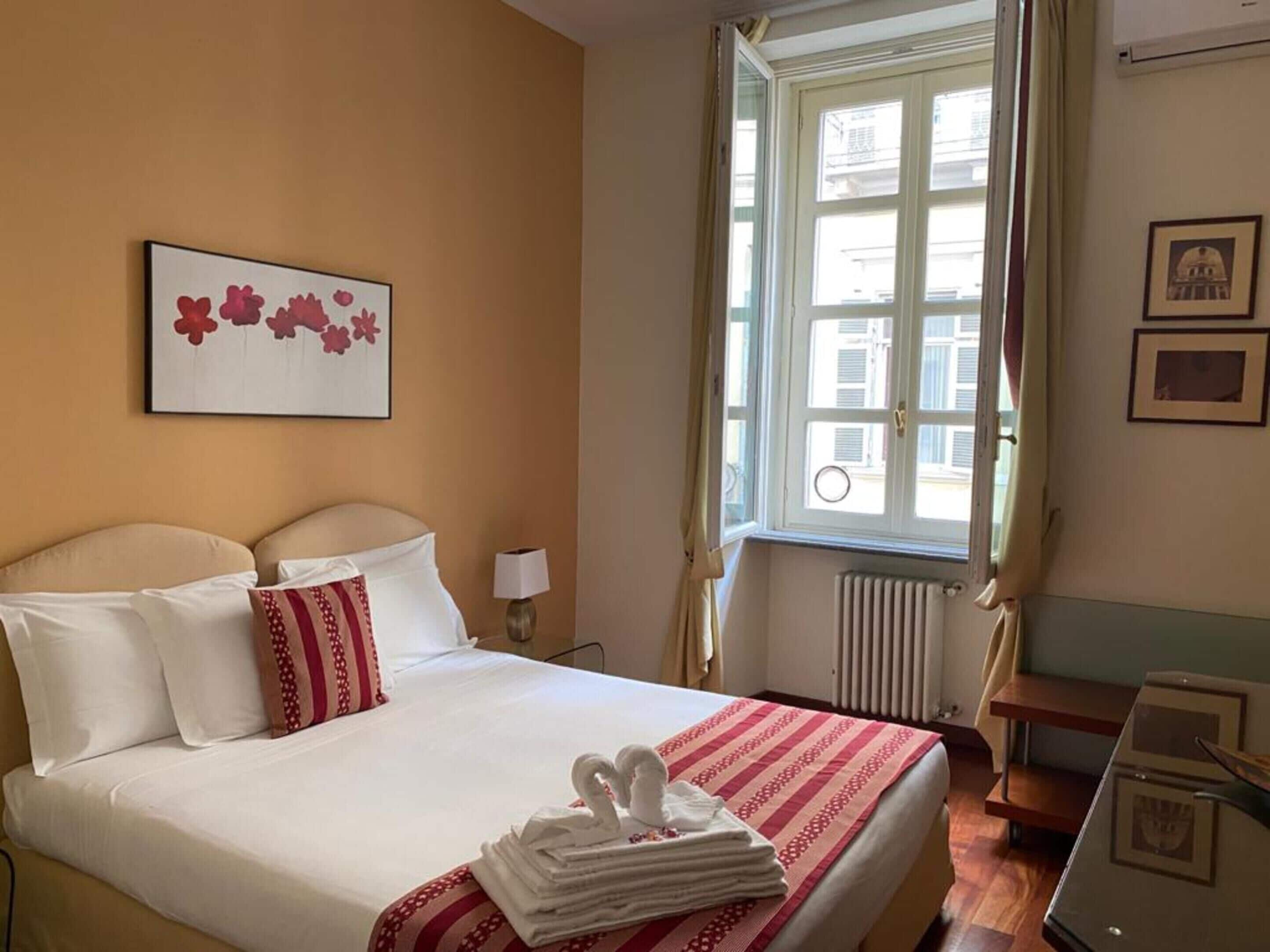 Habitación de hotel con cama, ventana con cortinas, radiador, escritorio y un cuadro en la pared.