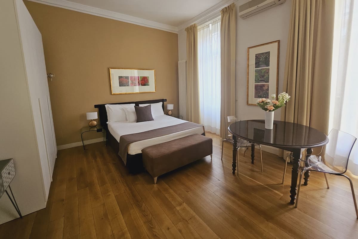A hotel room with a bed, bedside tables, a round dining table with chairs, a vase with flowers, and a framed picture on the wall.