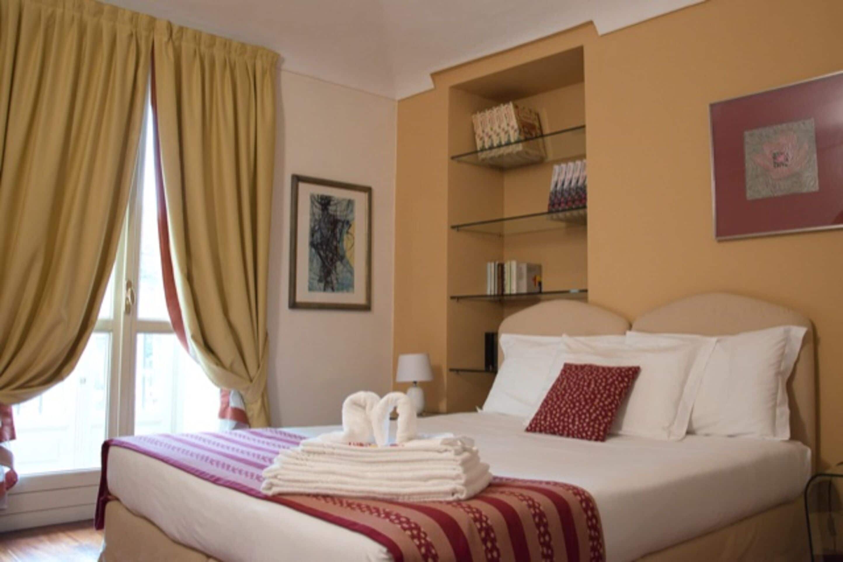 Una habitación de hotel con una cama, una mesita de noche, una lámpara, una ventana con cortinas, un estante con artículos de tocador y un cuadro en la pared.
