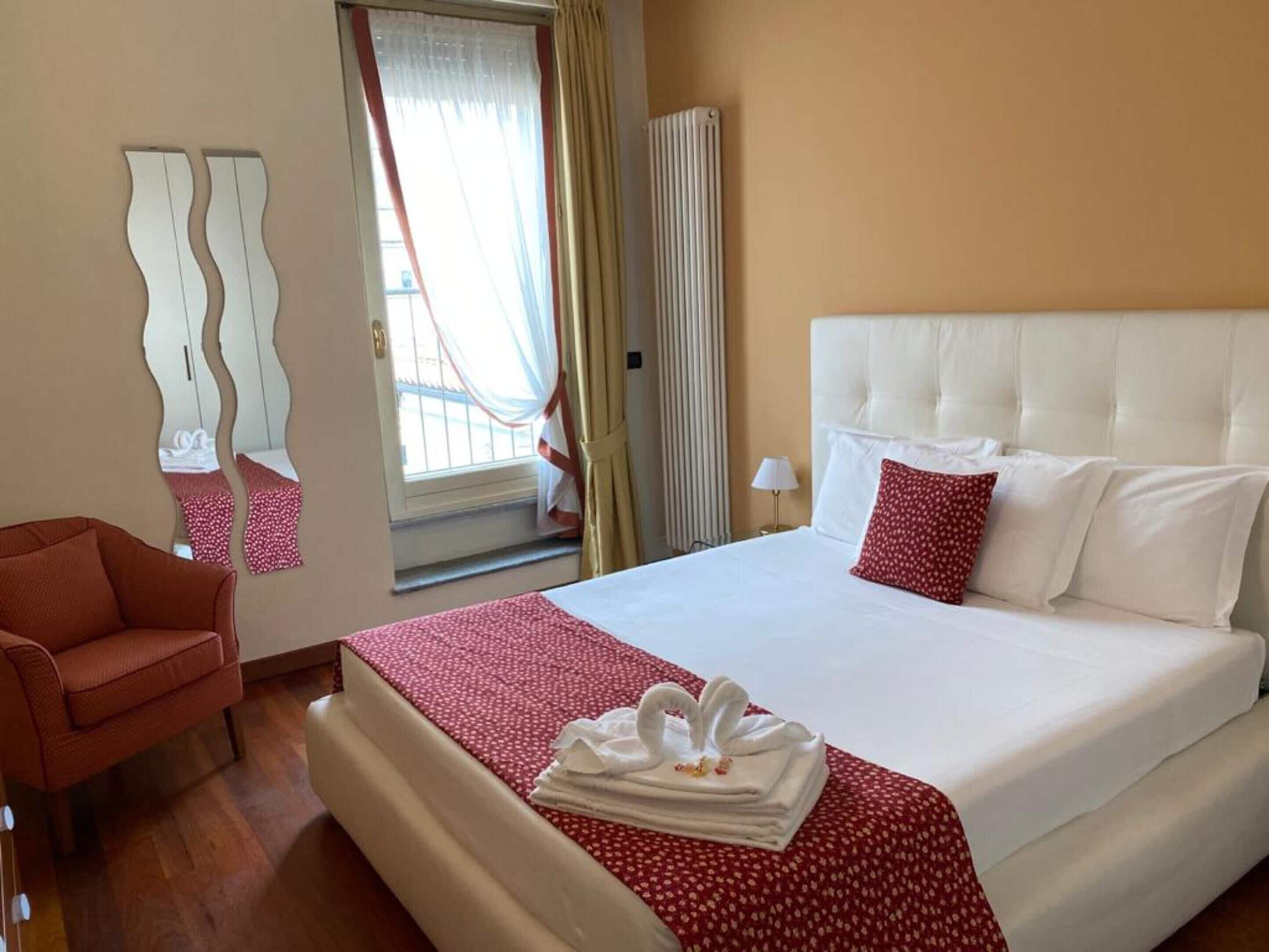 A hotel room with a large bed, a red and white patterned bedspread, a white headboard, a red pillow, a mirror with a wavy frame, a wooden floor, and a window with curtains.