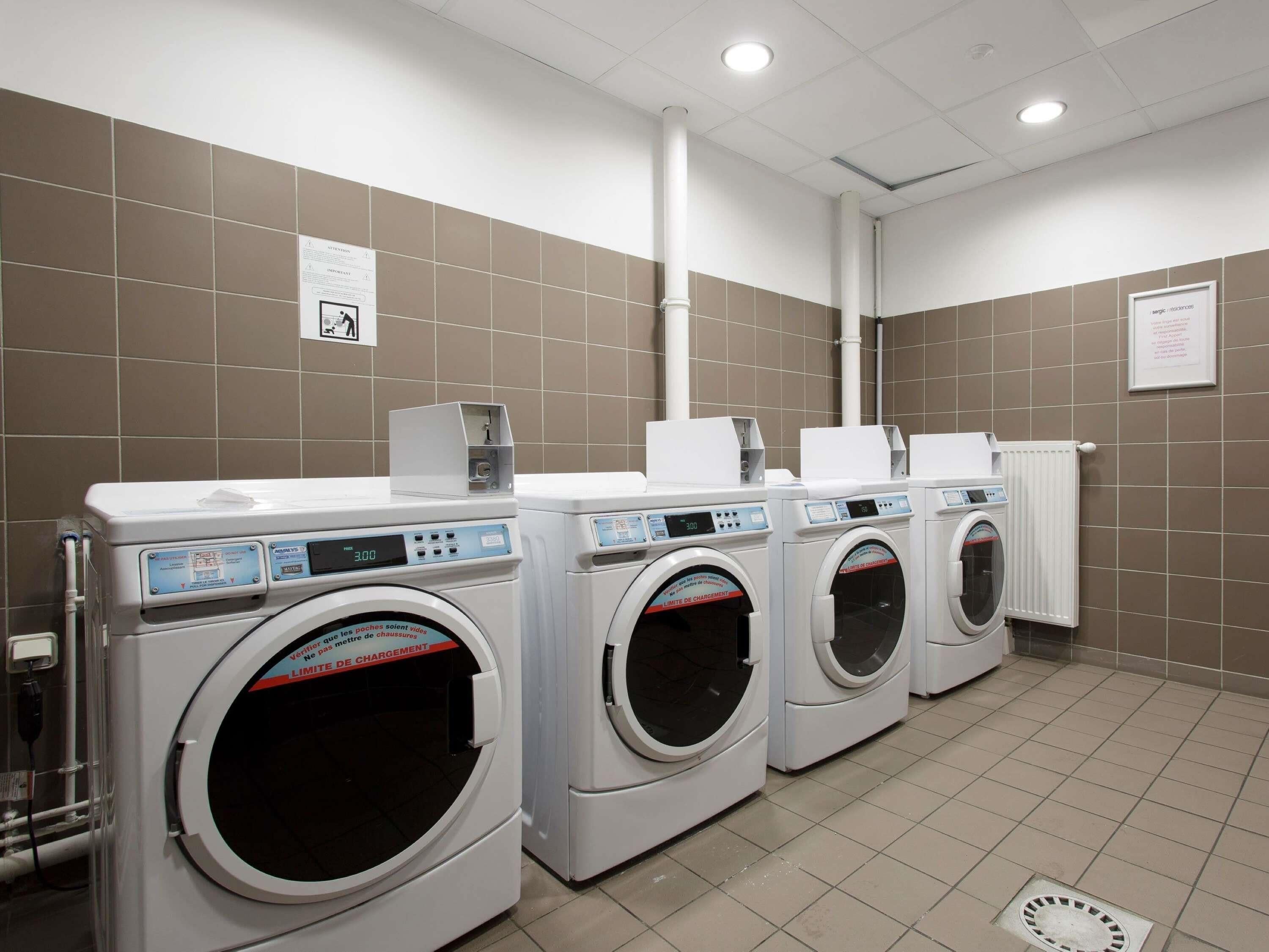 File di lavatrici bianche ad uso frontale in una lavanderia a gettoni con pareti piastrellate color beige e soffitto bianco.