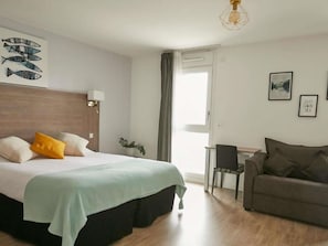 A hotel room with a bed, a desk, a sofa, a window, and artwork on the walls.