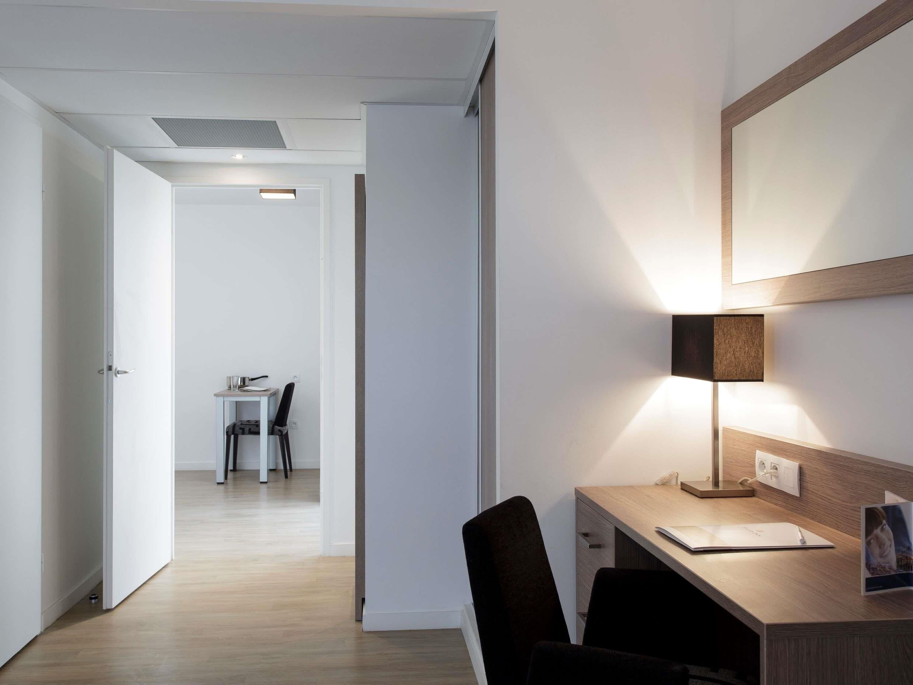A modern hotel room with a desk, chair, and a lamp. There is a door leading to another room and a hallway with a table and chairs.