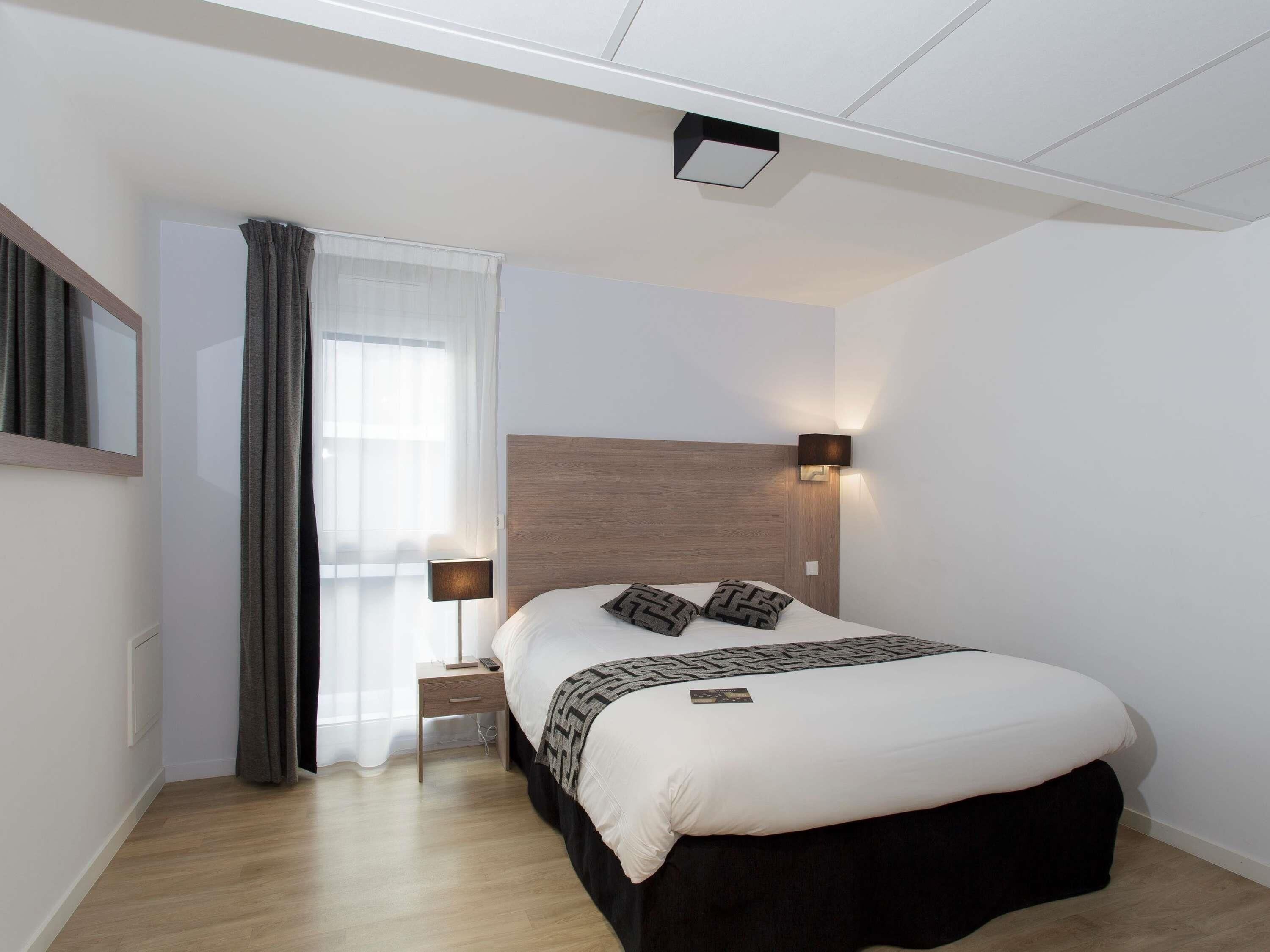 Una habitación de hotel moderna con una cama grande, lámparas de noche, una mesita auxiliar pequeña y un ventanal con cortinas.