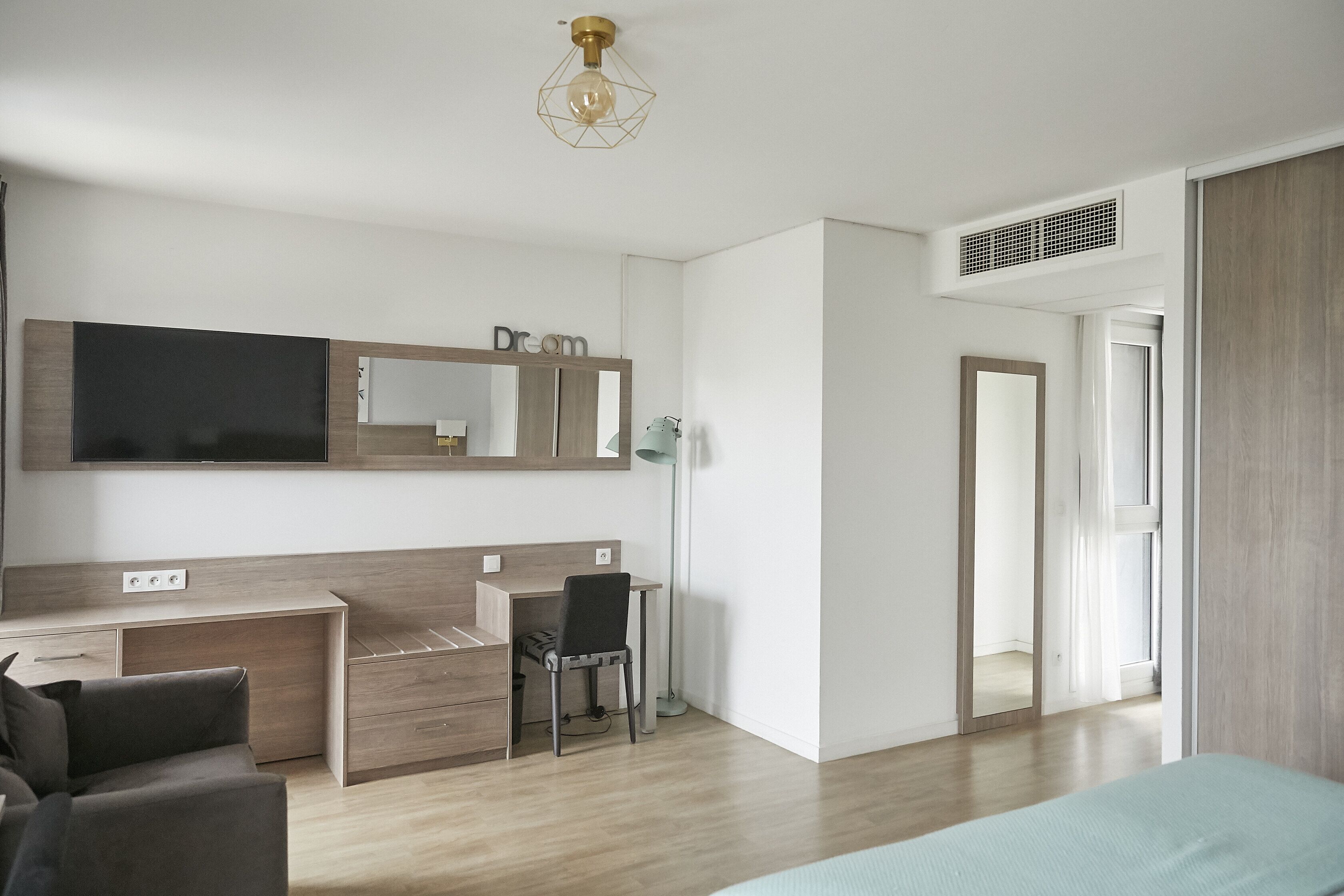 Une chambre moderne avec un lit, un bureau et une chaise. Un grand miroir est placé au-dessus du bureau, et un luminaire est fixé au plafond.