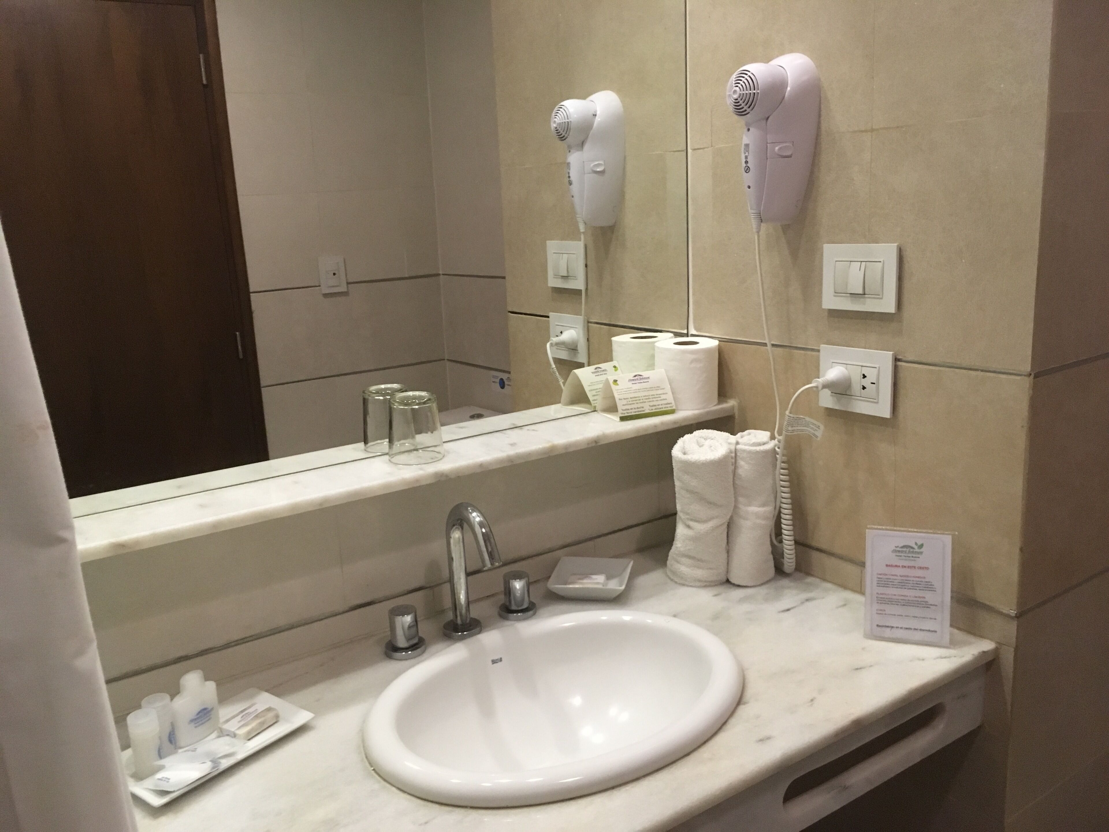 A hotel bathroom with a sink, mirror, hairdryer, and toiletries.