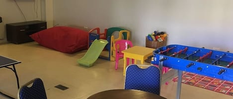 Un área de juegos infantiles con mesa de fútbol de mesa, sillas de colores, un tobogán pequeño y un televisor montado en la pared.