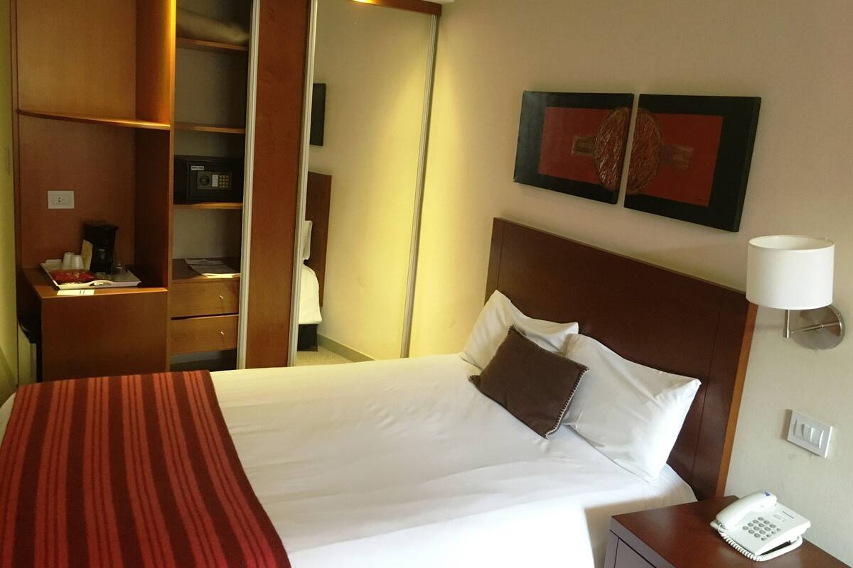 Habitación de hotel con una cama, una mesita de noche con teléfono, un espejo, un armario y dos cuadros en la pared.