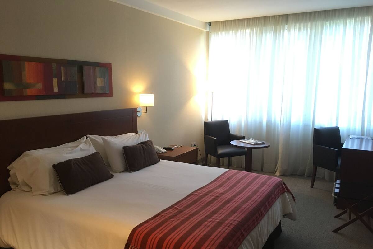 Una habitación de hotel con una cama grande, un escritorio, una silla, una lámpara y una ventana con cortinas.