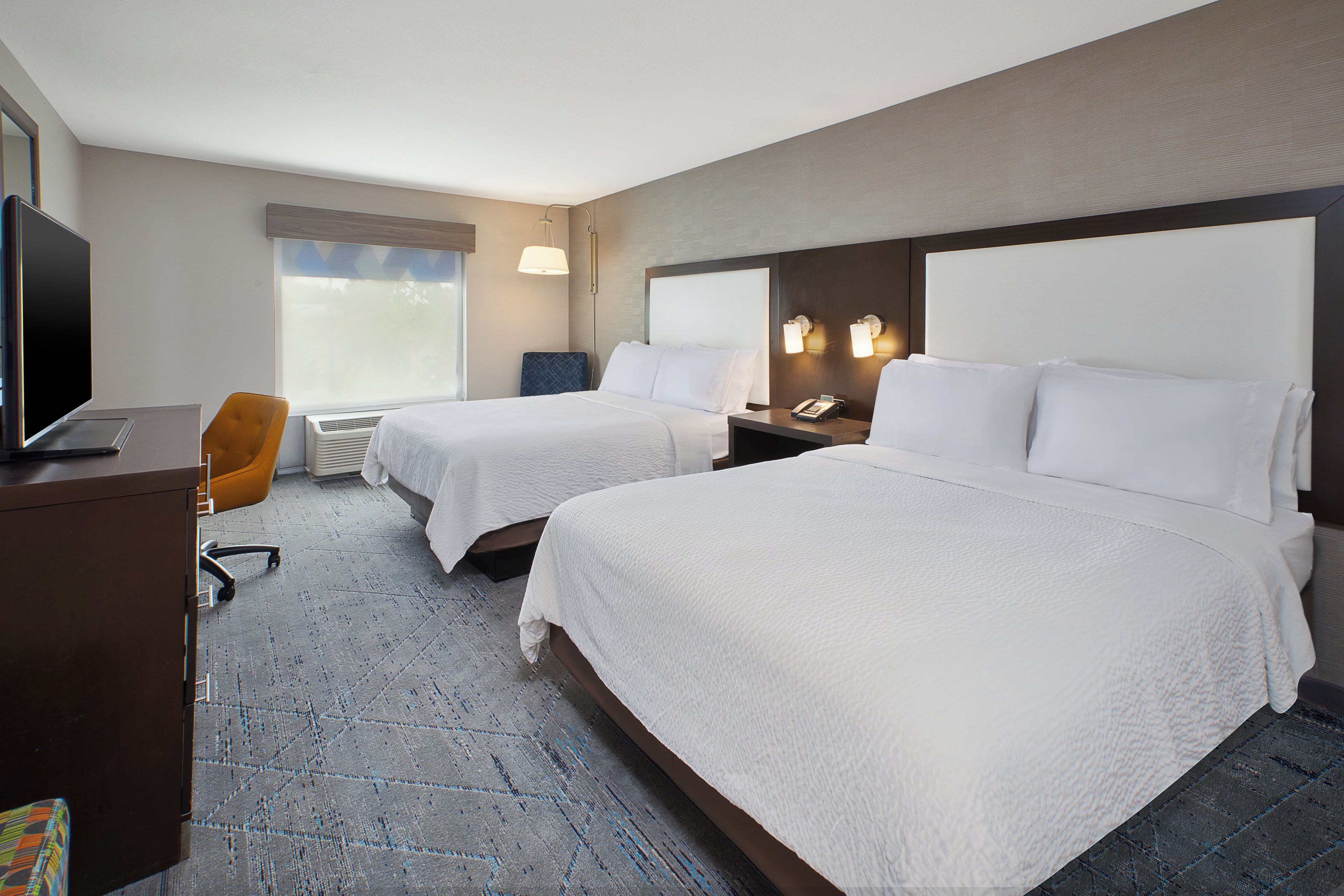 A hotel room with two beds, a desk with a television, a chair, and a window with blinds.