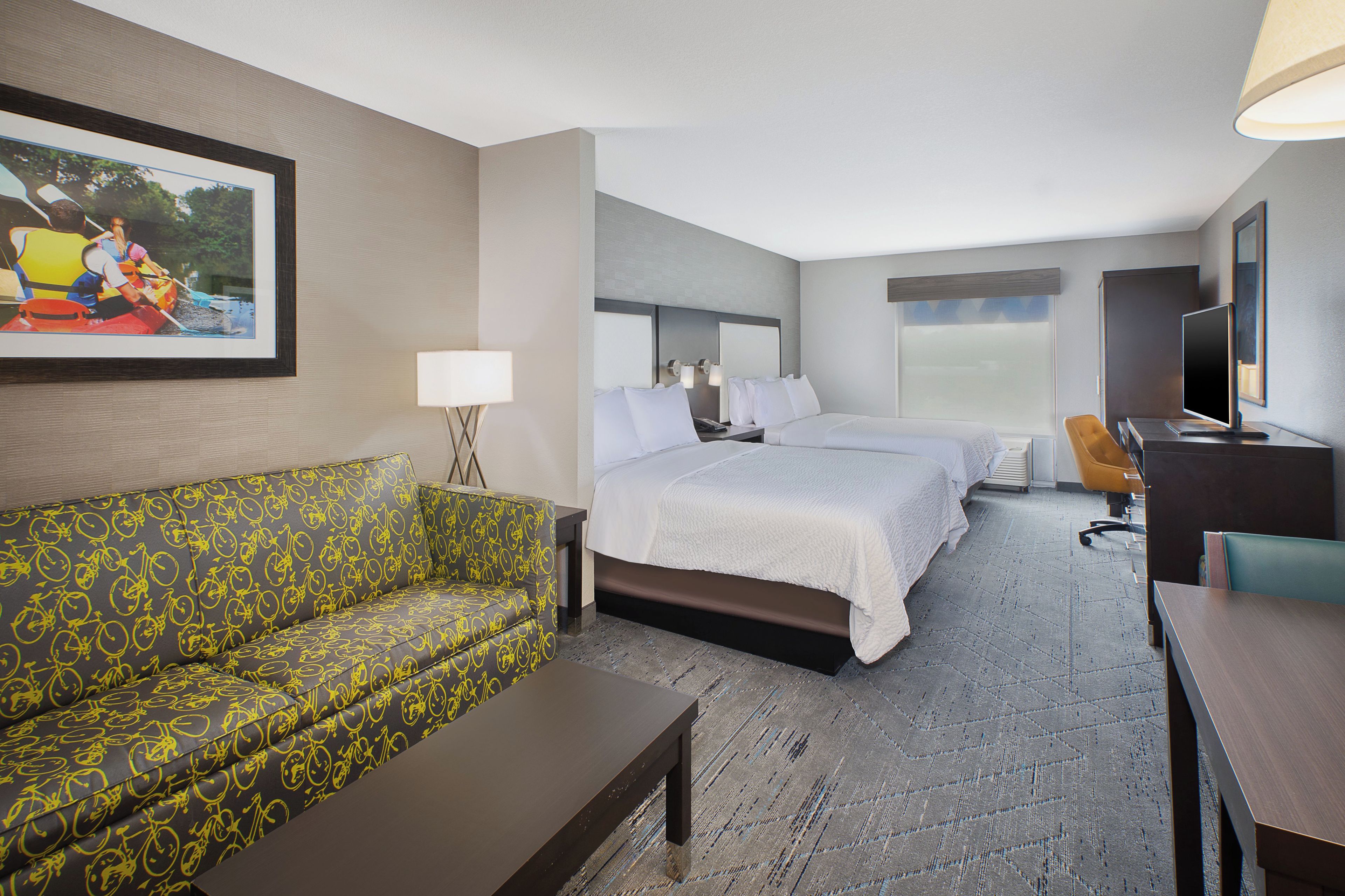 A hotel room with a bed, a sofa, a desk, a TV, and a painting on the wall.