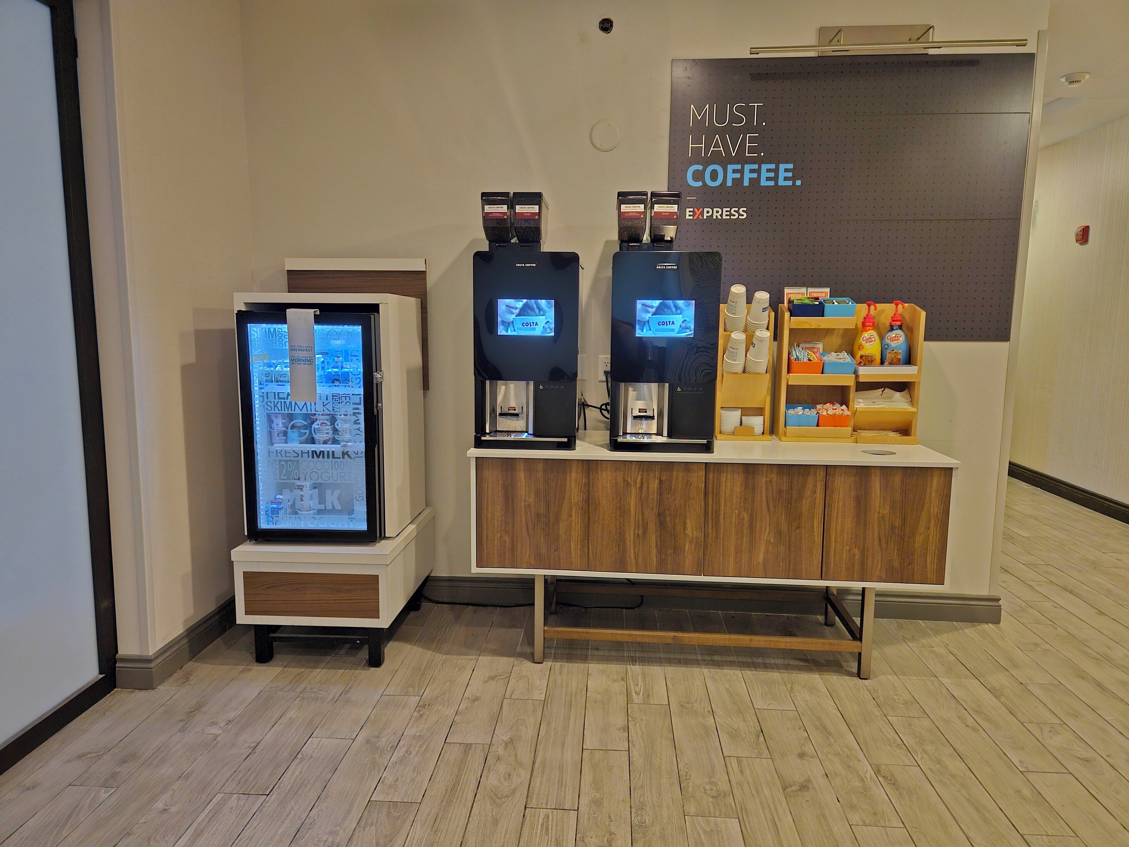 A coffee station with a digital display, a refrigerated display case, and a shelf with coffee cups and condiments.