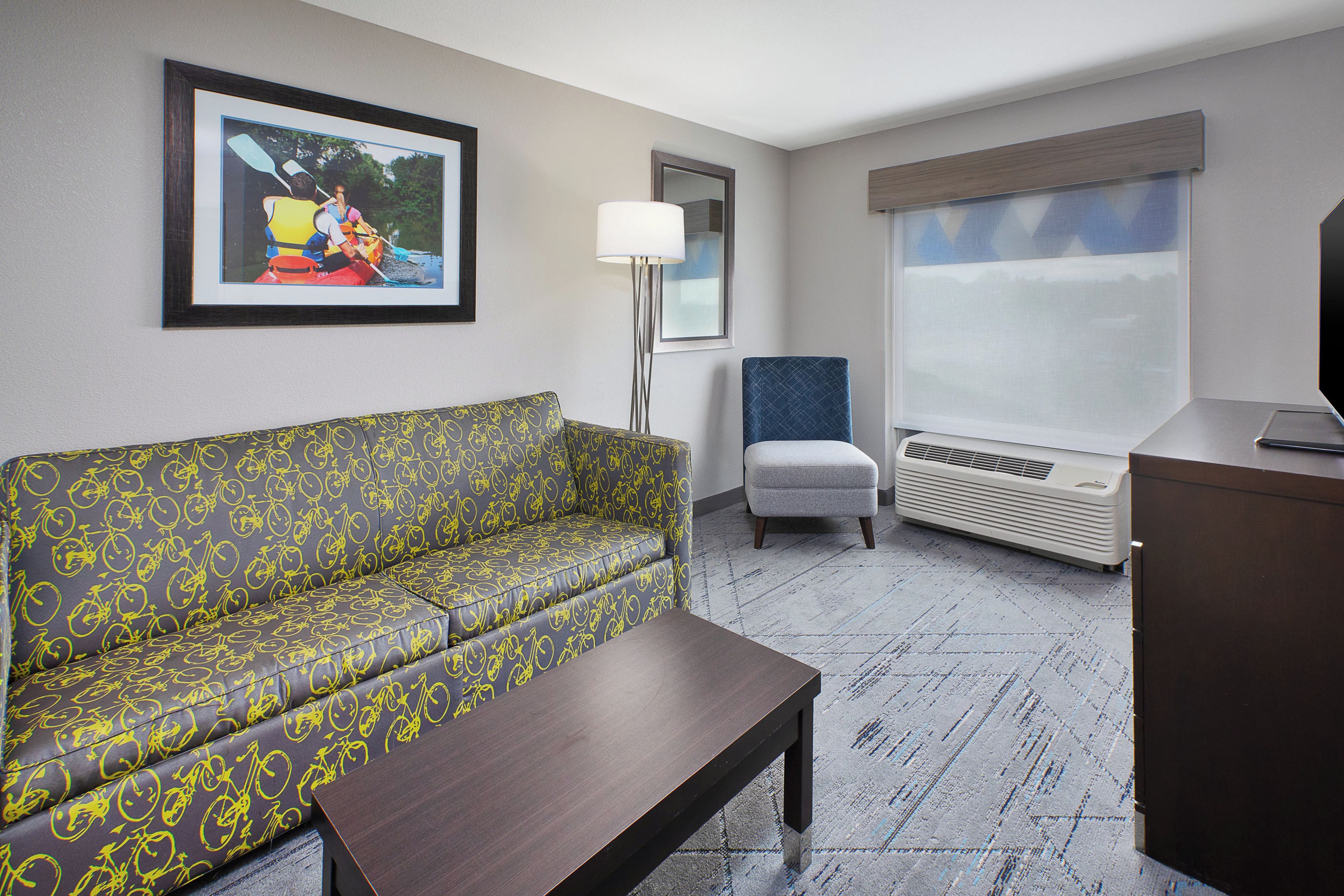 A hotel room with a sofa, a chair, a desk, a lamp, and a large window.