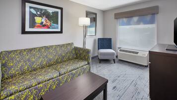 A hotel room with a sofa, a chair, a desk, a lamp, and a large window.