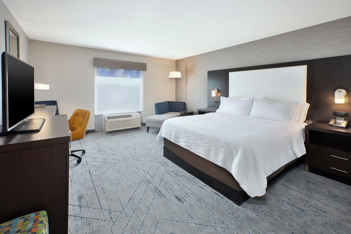 A hotel room with a bed, a desk with a computer, a chair, a window with blinds, and a wall-mounted TV.