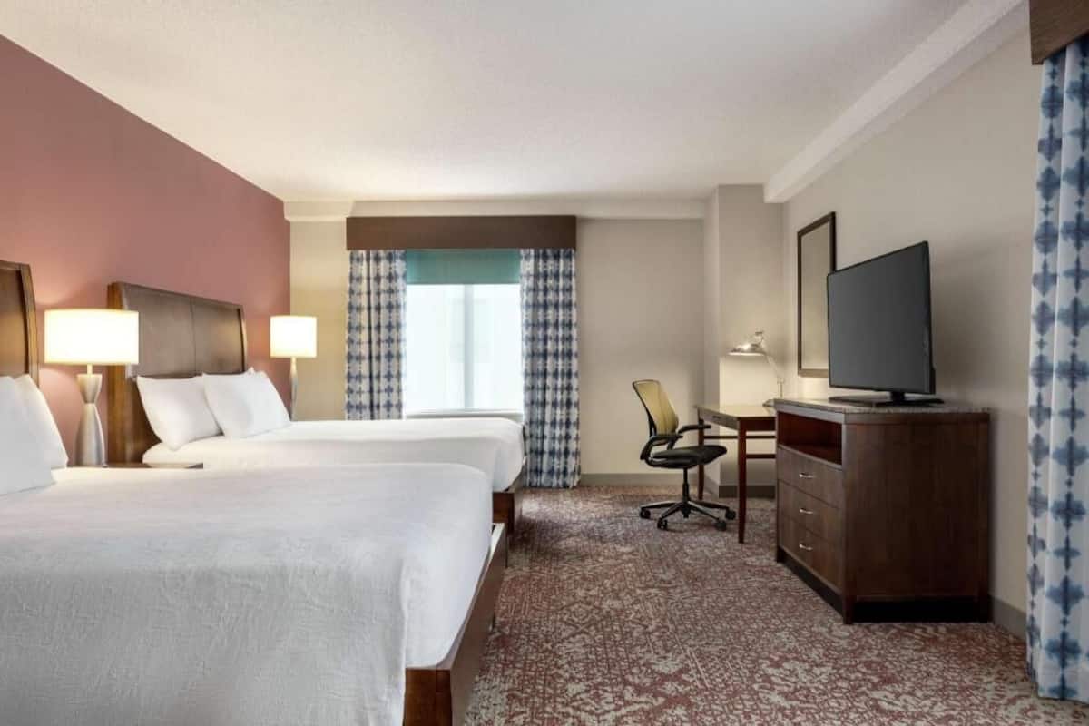 A hotel room with two beds, a desk, a television, and a window with patterned curtains.