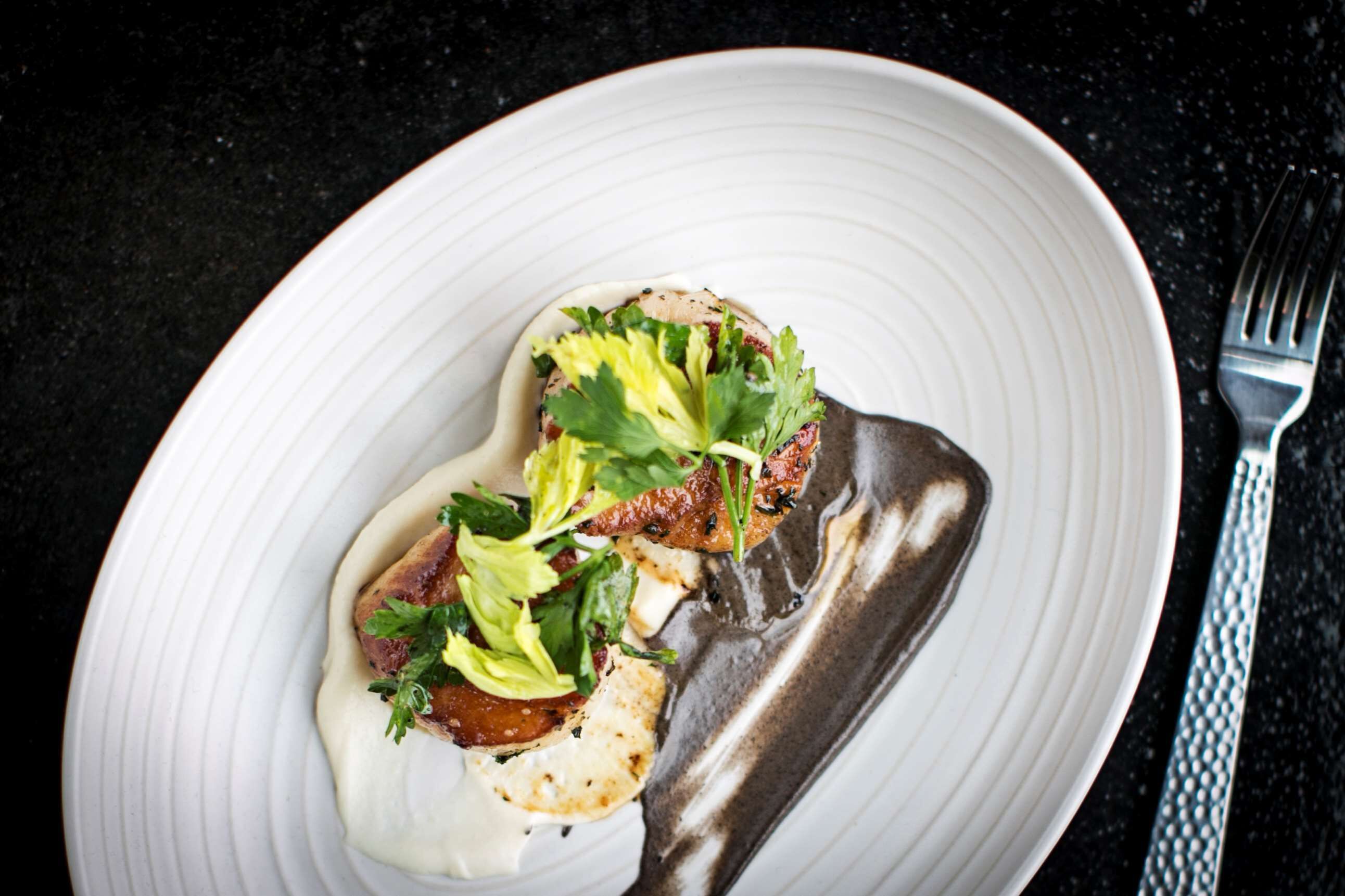 Une assiette blanche contenant un morceau de viande, des légumes verts et de la sauce, posée sur un plan de travail foncé.