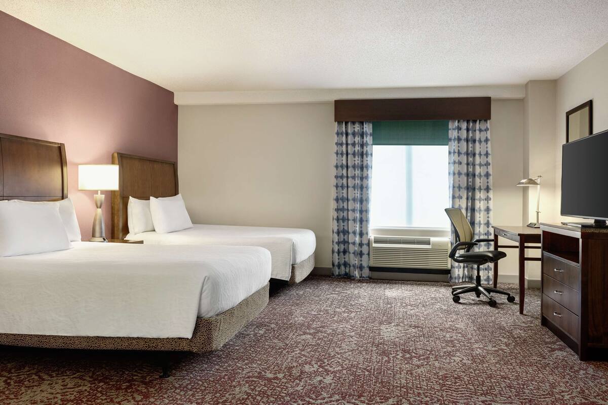 A hotel room with two beds, a desk, a chair, a TV, and an air conditioning unit.
