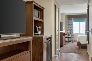 Habitación de hotel con escritorio de madera, televisor, cafetera, lámpara de escritorio, silla y cama.