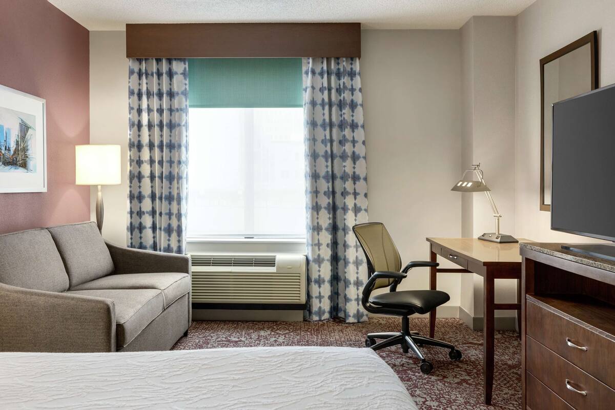 A hotel room with a bed, a sofa, a desk with a chair, a TV, and a window with curtains.
