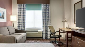 Habitación de hotel con cama, sofá, escritorio con silla, televisor y ventana con cortinas.