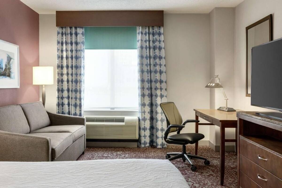 A hotel room with a bed, a desk with a chair, an air conditioner, a television, and a window with patterned curtains.