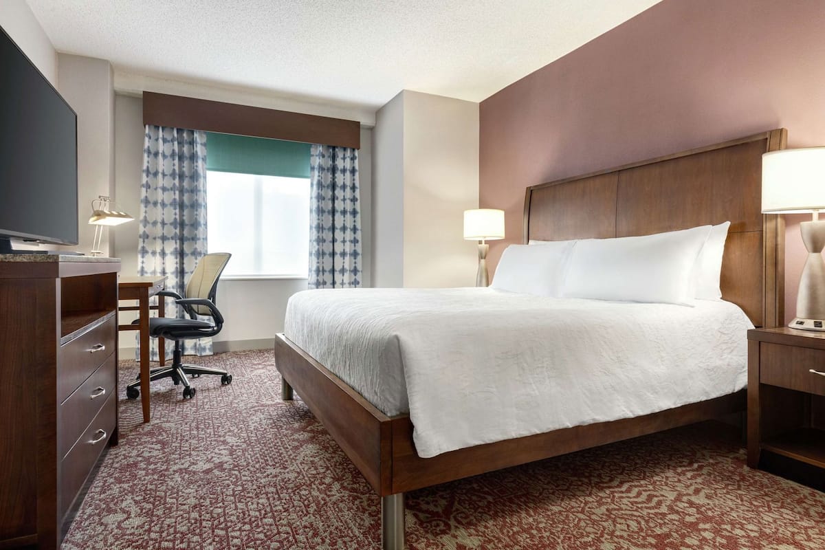 A hotel room with a bed, desk, chair, TV, and lamps.