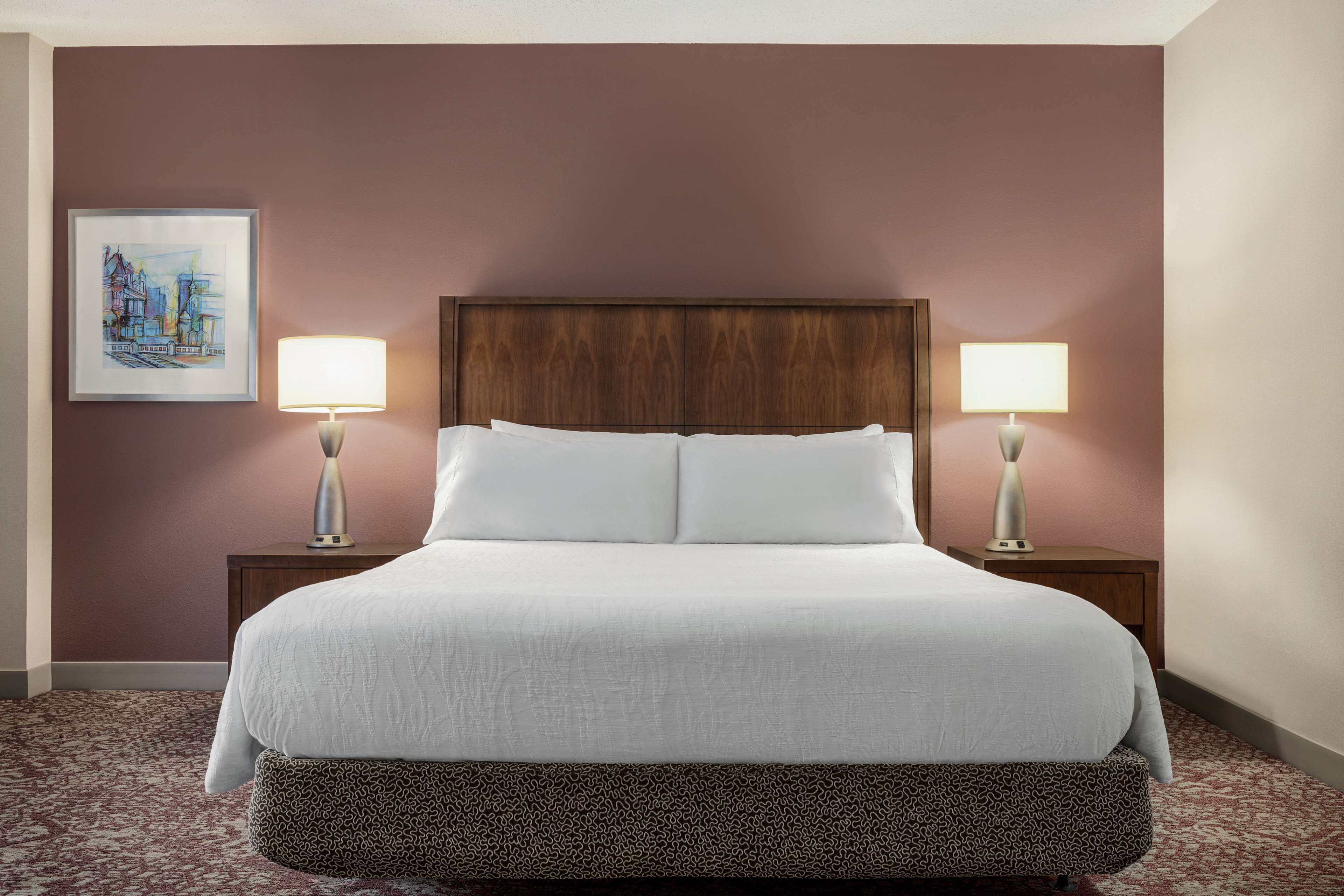 Een netjes opgemaakt bed met een houten hoofdbord, twee bedlampen en een ingelijste foto aan de muur.