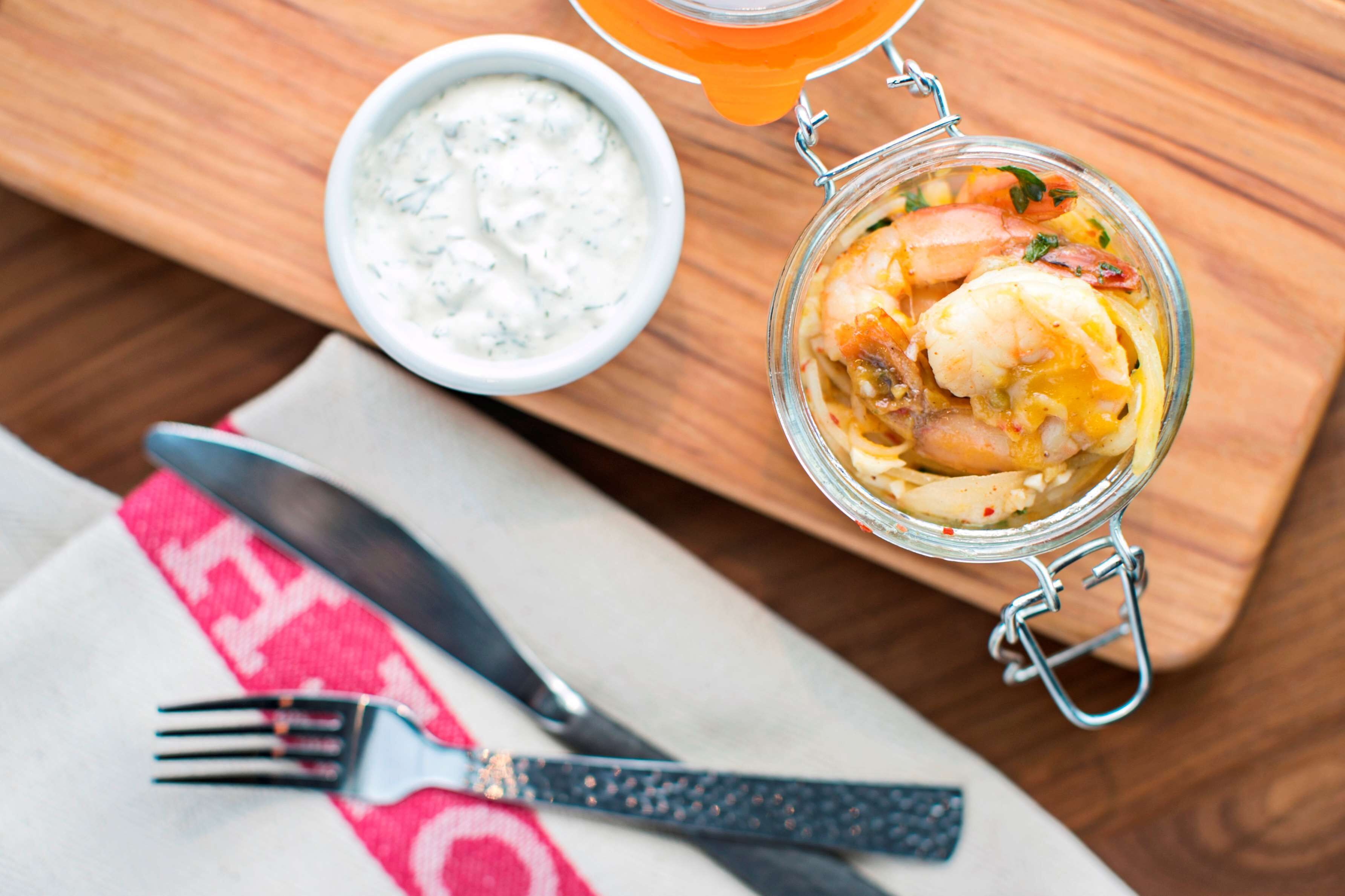 Un tarro de vidrio con cierre metálico que contiene un plato de mariscos, un cuenco pequeño de salsa blanca y un cuchillo y tenedor sobre una mesa de madera.
