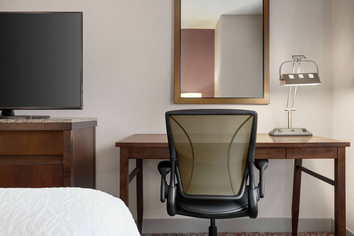 A hotel room with a bed, a desk with a chair, a television, a desk lamp, and a mirror.
