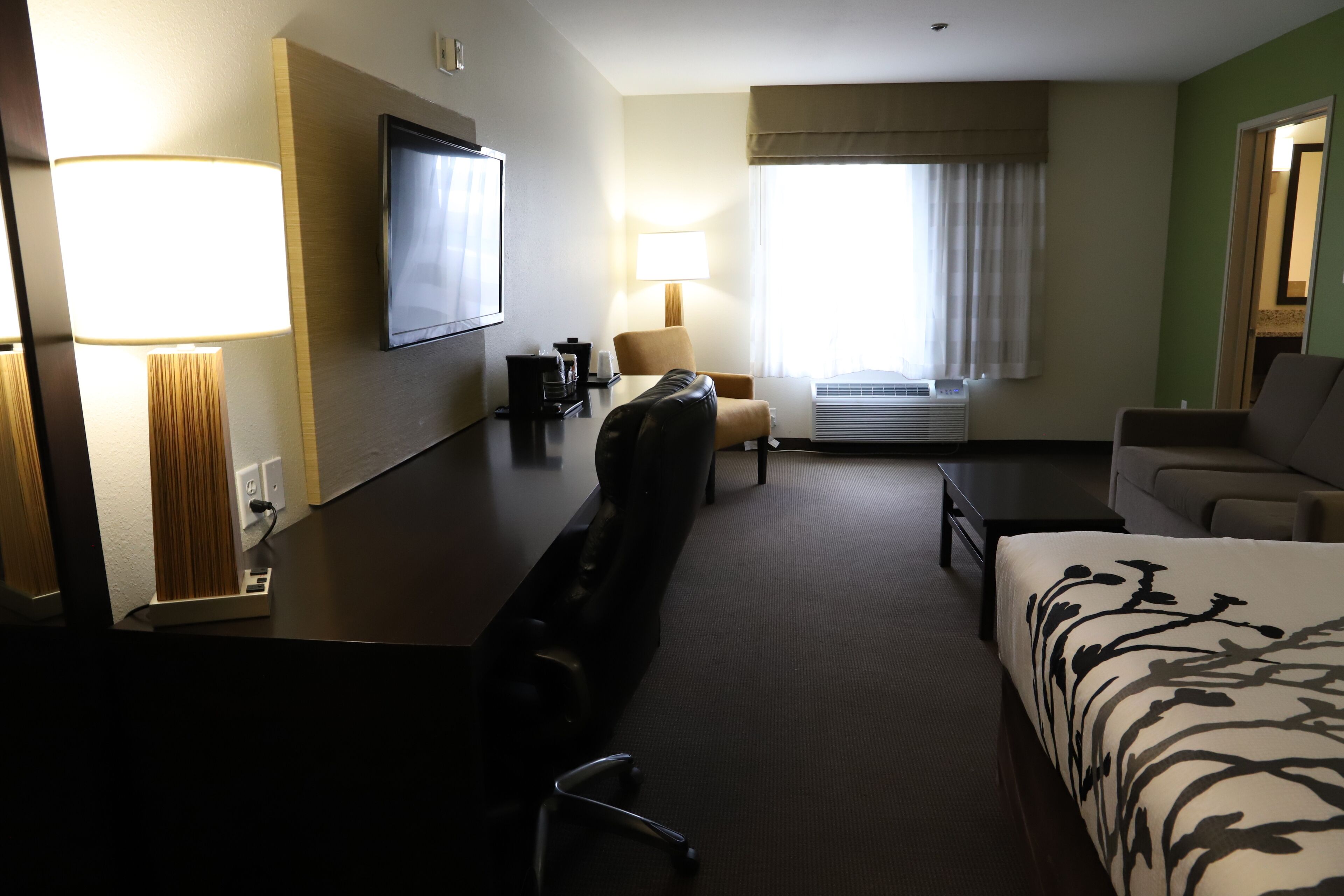 A hotel room with a bed, a sofa, a desk with a lamp, a television, and a window with curtains.