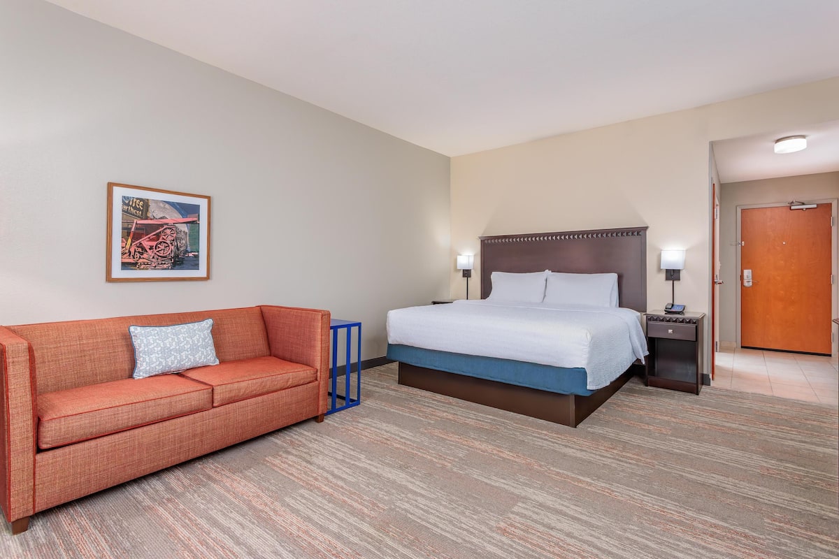 A hotel room with a bed, a sofa, a nightstand, a lamp, and a framed picture on the wall.