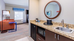 A modern kitchen with granite countertops, a stainless steel sink, and a microwave. There is a small refrigerator, a blue chair, and a window with blinds. - Hampton Inn & Suites Parkersburg Downtown (Parkersburg)