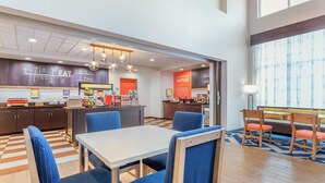 A modern dining area with a checkered floor, a central table with blue upholstered chairs, and a counter with a menu board. - Hampton Inn & Suites Parkersburg Downtown (Parkersburg)