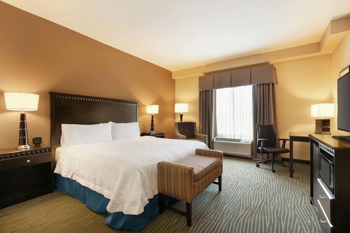 A hotel room with a large bed, two bedside tables with lamps, a desk with a chair, a television, and a window with curtains.