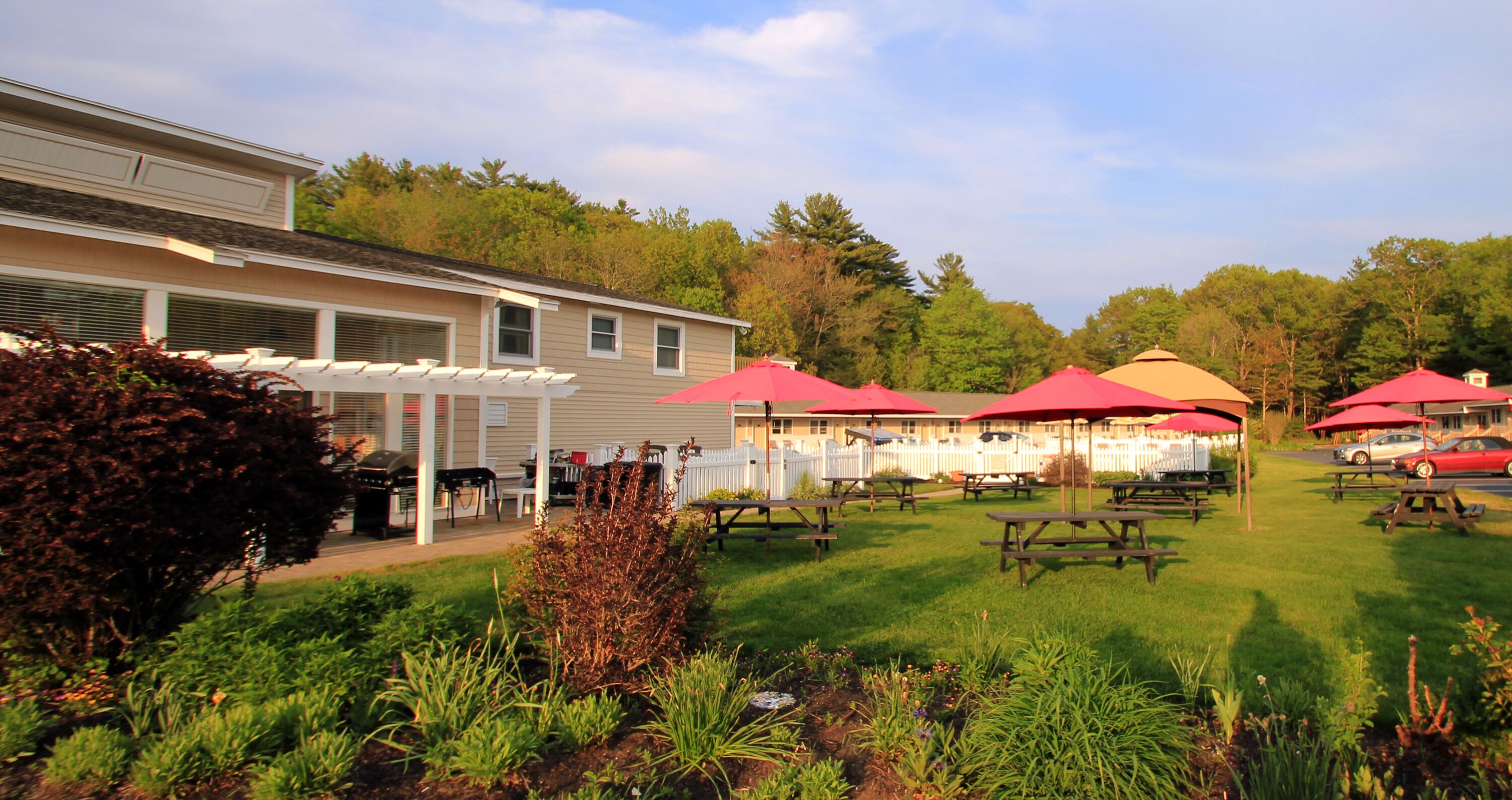Ein Garten mit Picknicktischen und roten Sonnenschirmen, umgeben von einem gepflegten Rasen und einem Haus.