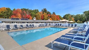 Une piscine rectangulaire bordée de chaises longues et d’ombrelles, entourée d’une clôture blanche en bois et d’arbres aux feuilles d’automne.