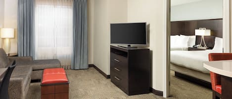 A hotel room with a bed, a desk, a television on a dresser, a chair, a sofa, and a window with curtains.