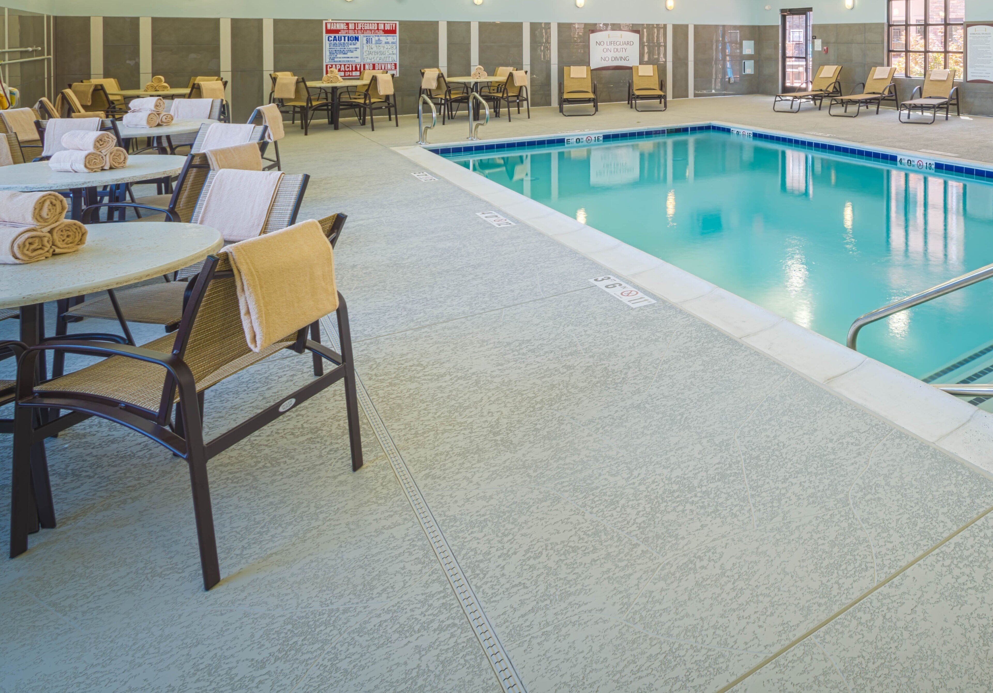 Área de piscina interna com espreguiçadeiras e mesas, e placas indicando ausência de salva-vidas e proibição de mergulho.