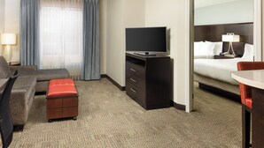 A hotel room with a sofa, a TV on a stand, a desk with a chair, a small table, a mirror, and a bed with a nightstand.